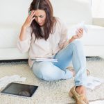 zmartwiona kobieta siedzi na kremowym dywanie oparta o białą sofę i liczy rachunki